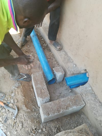 Two individuals are working on a construction or repair project. Blue pipes are being laid in a trench dug alongside a wall, with sections visible entering an opening in the wall. Several rectangular blocks or bricks lie nearby, along with a hammer. The ground is composed of soil and loose debris, and the workers are dressed casually, wearing sandals. The scene suggests a simple plumbing or drainage setup.