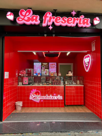 The image shows the storefront of "La Freseria" or "La Maduixeria," a dessert shop, likely specializing in strawberry-themed treats. The shop, located in Granollers, Spain, based on other cues, has a bright red interior with a tiled design. There are display cases filled with desserts, digital menus showing pictures of items such as "Fresalad" and "Celat de Nata," and neon signs. One sign displays "La freseria" vertically outside the store, while inside a heart-shaped neon sign reads "FRESAME HUCHO". A person is partially visible behind the counter. A trash can is located near the entrance.