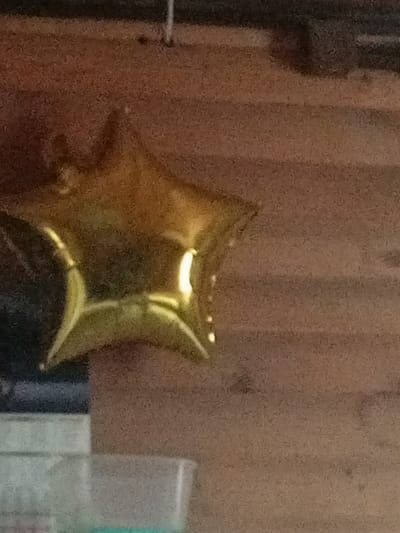 A gold star-shaped balloon is suspended from a wire against a textured wooden wall. The balloon is inflated and reflects light, with a bright spot visible on its surface. Below the balloon, a portion of a clear plastic container is visible. The image is grainy and appears to have been taken in low light conditions. The location is identified as Засопка, Russia, though specific details of the urban environment are not discernible from the image.