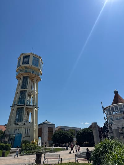 The image shows the Siófok Water Tower in Hungary under a clear, blue sky. A bright sunlight streak is visible across the upper left. The tower is the primary subject, showing its octagonal structure with multiple levels of glass-enclosed observation decks. The tower's support structure is painted in a cream color. To the right of the water tower, a section of a pedestrian walkway is visible. People are walking and standing along the walkway. Trees and other buildings are also partially visible in the background.