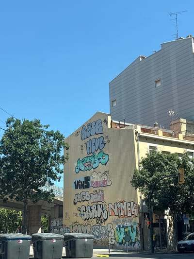 The image shows a building covered in graffiti, located in Girona, Spain. The building is tall and painted a light yellow color. The graffiti is varied in color and style, with large bubble letters and smaller tags. Some of the visible words include "NEVE", "VINS", "SARK", and "NIMEK". The building is situated in an urban environment with trees, trash bins, a traffic light, and a modern grey building in the background. A portion of an overpass is also visible. The sky is clear and blue.
