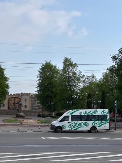 The image depicts a white van with green lettering that reads "Габария" parked on a city street in Akhieli, Georgia. The vehicle is situated adjacent to a pedestrian crosswalk. Behind the van, there is a park or landscaped area featuring a statue in front of a medieval-style structure made of light-colored brick or stone. Trees surround the area, and overhead power lines are visible. The sky is overcast. There are pigeons in the street.