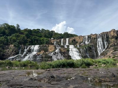 The image displays the Pongour Waterfall in Tân Phú, Vietnam, under a partly cloudy sky. The waterfall cascades down a series of rocky terraces, creating a wide, multi-tiered effect. Lush green vegetation covers the cliffs above and surrounds the base of the waterfall, where a calm body of water reflects the scene. In the foreground, dark, wet rocks with shallow pools of water are visible. The image is a landscape shot, emphasizing the natural beauty and scale of the waterfall.