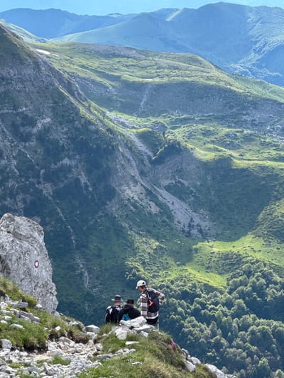 In Jošanica, Montenegro, on a steep, rocky mountainside, a group of three hikers stand near the edge of a cliff. The person in the foreground, wearing a plaid shirt and a white baseball cap, faces away from the camera. To their left, two other individuals are also facing away, one wearing a hat and the other a black hoodie with a blue patterned backpack. The mountain range behind them features lush green vegetation, with a rocky, eroded cliff face on the left and rolling hills covered in grass and trees extending into the distance under a clear blue sky. A red and white trail marker is visible on a large rock in the foreground.