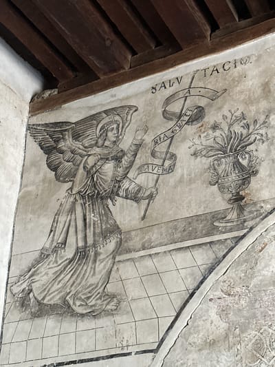 This image depicts a fresco on a wall, likely within the Augustinian Convent of San Miguel in Huejotzingo, Mexico. The fresco features an angel in flight, rendered in grayscale. The angel has large, feathered wings and is adorned with a halo and a jeweled crown. It is dressed in a long, flowing robe and holds a staff with a banner attached, inscribed with Latin text. The banner reads "AVE MARIA GRATIA PLENA" on one side and a partial "SALVATOR" on the other. To the right of the angel, a decorative vase filled with flowers is depicted. The background is textured, with a tiled floor pattern beneath the angel. The upper portion of the image shows wooden beams of a ceiling, suggesting the fresco is located indoors. The overall style is reminiscent of Renaissance or Baroque religious art.