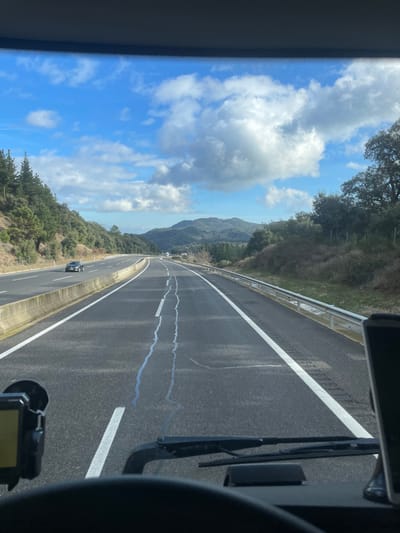 The image captures a view from inside a moving vehicle, likely a bus or truck, looking forward and slightly to the right onto a multi-lane highway. The scene depicts a sunny day with clear blue skies dotted with scattered white, fluffy clouds.
The central subject is the asphalt highway, featuring distinct white lane markings, including solid lines separating lanes and a dashed line on the shoulder. Visible on the road surface are several long, wavy patches of grey asphalt repair. A metallic guardrail runs along the right edge of the highway, separating it from the surrounding terrain.
On both sides of the highway, the landscape is characterized by rolling hills or mountains densely covered with green trees and vegetation, suggesting a forested environment. Further in the distance, more mountains are visible on the horizon. A small, dark-colored car can be seen on the opposite lanes of the highway, moving away from the viewer.
Within the vehicle's interior, visible on the right side of the frame, are parts of the dashboard, windshield wiper blades, and what appears to be a mounted device or camera.
The activity taking place is travel along a highway, likely through a scenic, rural or semi-rural region. Given the location context of Arbúcies, Spain, this aligns with the mountainous and forested landscape characteristic of Catalonia. The time of day appears to be daytime with bright sunlight, indicating pleasant weather conditions for driving. No visible text is clearly legible in the image.