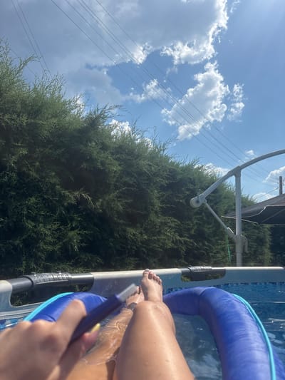 The image shows a close-up of a person's legs and feet as they sit in a swimming pool. The person is holding a blue pool cleaning tool. The pool has an inflatable blue ring around its edge. Sunlight glistens on the water and the person's wet skin. In the background, a dense green hedge or tree is visible, and beyond that, a bright blue sky with fluffy white clouds. Power lines stretch across the sky. The scene suggests a sunny day, likely in the late morning or early afternoon, given the bright light. The setting appears to be a backyard or outdoor recreational area in Viden, Bulgaria, possibly during the summer. There is no visible text in the image.