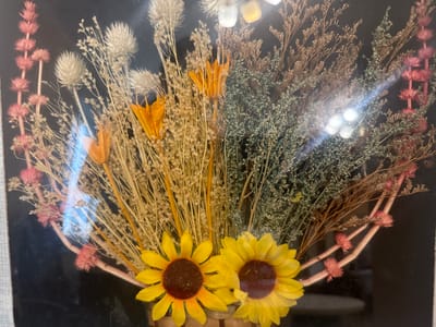 This image shows a framed arrangement of dried flowers, including sunflowers, with a dark background. The arrangement appears to be a decorative piece, possibly a wall hanging or a centerpiece. The flowers are varied in color and texture, with yellow sunflowers at the bottom, flanked by lighter-colored dried grasses and twigs. There are also pink and orange accents within the arrangement. The image is taken at a slight angle, with some glare from light sources visible on the surface of the frame. The location in the city of Kuril'sk, Russia, is not determinable from the visual information present in the image. There are no people, actions, or interactions depicted. The environment appears to be indoors, with the dark background suggesting the absence of natural light or a deliberate stylistic choice.