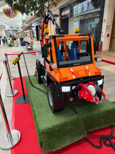 A large-scale LEGO model of an orange and black Unimog utility truck is prominently displayed within a modern shopping center in Mont-Saint-Martin, France. The detailed model features an articulated crane arm with red and black components at the rear, two yellow warning beacons mounted on the cab roof, and a gray utility bed. Its front is equipped with illuminated white headlights and a red, white, and gray front attachment, possibly a plow or shovel. Blue LEGO bricks form the interior seating. The "UNIMOG" designation is visible on the front grille area.

The LEGO truck rests on two elevated, interconnected platforms covered in artificial green grass, which are placed atop a red carpet. A perimeter around the display is defined by silver stanchions connected by red ropes. A black stand within this roped-off area holds a yellow remote control, connected to the truck by a coiled black cable. Additional coiled black cables extend from the truck's front and rear, disappearing beneath the grass platforms.

The background shows a brightly lit indoor mall environment with a light-colored tiled floor and a high glass ceiling. Multiple storefronts are visible, including one labeled "FOOT KORNER" with a partially lowered security gate. Potted trees line the walkway. In the middle distance, several individuals are present, some observing children operating small ride-on toy vehicles, one green and one blue, along the mall's pedestrian path. Benches and other public seating are also discernible.