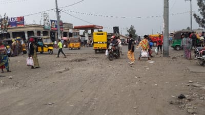 This image depicts a bustling street scene in the city of Rukoko, Democratic Republic of the Congo. The foreground consists of an unpaved, dusty, and uneven ground surface with scattered debris.
In the midground and background, numerous individuals, vehicles, and structures are visible under an overcast sky. People, both male and female, of varying ages, are distributed across the scene. Many are dressed in colorful, patterned fabrics. On the left, a woman wearing a black top and a patterned skirt stands facing right, holding a white and red striped bag. A young child in a blue and yellow patterned outfit is visible to her left.
Vehicles present include multiple yellow auto-rickshaws (tuk-tuks), several motorcycles, and a distinctive yellow box-shaped vehicle, possibly an armored or utility truck, positioned in the center-right. One green auto-rickshaw is visible on the far right. Pedestrians are seen walking, standing, and interacting, some appearing to be engaged in commerce or transit.
In the background, various buildings line the street. On the left, commercial structures display large advertisements, including a prominent "FLEXBET" sign. In the center-right background, a red and white canopy of a gas station is visible. Utility poles and a complex network of overhead power lines crisscross the upper portion of the image. The overall environment suggests an active urban or commercial area.