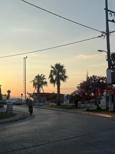 This image captures a sunset scene in Prinos, Greece. The sky is a gradient of soft yellow and orange, indicating the time of day. In the foreground, a power line stretches diagonally across the frame, with a wooden pole and wires. To the right, a paved road curves gently, bordered by a sidewalk with benches and some greenery. A person is riding a bicycle on the road, suggesting a leisurely activity.

Further back, palm trees with their fronds swaying add to the tropical feel of the location. Behind the trees, a red building is visible, possibly a structure associated with a waterfront or a local business. A tall tower with a metal frame stands on the left side of the road, possibly a lighting tower. In the background, hints of a ship or boat structure can be seen, along with more trees and buildings. The overall atmosphere appears calm and serene, typical of a coastal town at dusk. There is some visible text on a red sign that reads "PANAGIA THA".