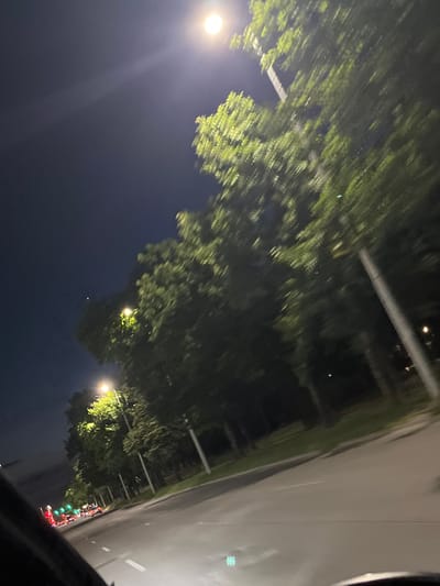 The image shows a street scene at night in Plovdiv, Bulgaria. A street light illuminates a row of trees lining the road. Cars are visible on the road, with their headlights and taillights creating streaks of light due to motion blur. The sky is dark, indicating nighttime. The perspective suggests the photo was taken from inside a moving vehicle.