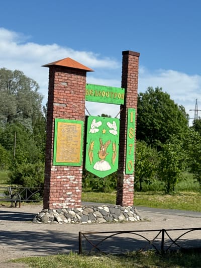 This image depicts a monument situated in Beloostrov, Russia, under a clear blue sky with sparse white clouds. The monument consists of two vertical brick columns with irregular stone bases. The left column is topped with a rust-colored pyramidal cap, while the right column has a flat, dark brown cap.

A green horizontal sign, bearing the Cyrillic text "БЕЛООСТРОВ" (Beloostrov) in yellow capital letters, is suspended between the two columns by chains. Below this, a large green shield-shaped sign, bordered in gold, hangs centrally. This shield features white stylized clouds, a golden five-pointed star, a golden bird resembling a goose or swan on a white mound, and golden wheat stalks. To the left of this central shield, a rectangular green-framed panel with illegible golden text is mounted on the left brick column. Vertically displayed on the right brick column are the golden numbers "1870".

The background is dominated by lush green deciduous trees and bushes. A paved pathway or road is visible in the foreground, with sections of grass and dirt. A wooden park bench and a section of decorative metal fence are present to the left of the monument. In the immediate foreground, a low, ornate metal fence partially frames the scene. In the far right background, a metal power line tower is discernible above the tree line. The scene is brightly lit by sunlight, casting distinct shadows on the pathway. No people or active interactions are visible in the image.