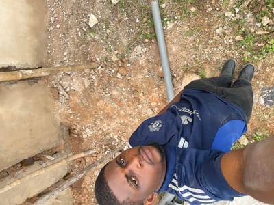 The image depicts a young man, appearing to be of Nigerian descent, standing outdoors in a possibly unfinished construction area. He is wearing a blue athletic shirt with white stripes and black pants, along with black boots. The ground is a mixture of dirt, gravel, and scattered rocks, with some sparse green vegetation. In the background, there are bamboo poles and a grey pipe, suggesting ongoing construction or repair work. The man's gaze is directed towards the camera. The scene is consistent with an outdoor setting in Okinano, Nigeria, given the visual cues of the environment and the appearance of the individual.