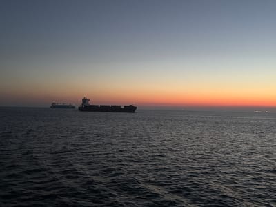 The image shows a wide view of a dark, choppy sea under a gradient sky of pale blue and warm orange, typical of a sunset or sunrise. Two large cargo ships are visible on the horizon. The ship closer to the viewer, on the right, is a dark silhouette against the lighter sky, showing a structured deck with lights. Further to the left, a second, smaller cargo ship is also visible, appearing as a dark shape against the fading light. The sea is filled with small waves. In the far distance, along the horizon, a faint line of lights suggests a distant shoreline or another group of ships. The scene is serene and maritime. The location of Fehmarn, Germany is not visually discernible from the image itself.