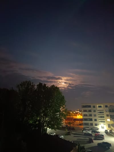 This is a nighttime, slightly low-angle, long shot capturing a scene in Târgu Jiu, Romania. The sky is a deep blue, transitioning to darker tones, with a prominent moon partially obscured by scattered clouds. A few stars are visible in the upper left portion of the frame. The moon is casting a soft, ethereal light.

In the foreground, a large, dark tree with lush green foliage occupies the left side of the image, its branches silhouetted against the sky and illuminated by unseen light sources. Beyond the tree, a paved parking lot is filled with several cars. The cars are of various models and colors, including white, silver, black, and red. Some cars are parked neatly, while others are at slight angles.

To the right of the parking lot, a multi-story modern building with a white facade and numerous windows stands prominently. Some windows are illuminated with warm yellow light, suggesting occupants inside. The building's design is contemporary, featuring clean lines and multiple balconies. In the background, other buildings and streetlights are visible, indicating an urban or suburban environment within Târgu Jiu. The overall atmosphere is calm and quiet, characteristic of a residential area at night. There are no people visible in the image.