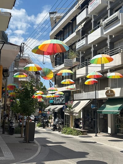 The image depicts a sunlit street in Giannitsa, Greece, characterized by numerous multi-colored, rainbow-striped umbrellas suspended high above the thoroughfare. The sky overhead is partly cloudy with significant blue areas.

The street, paved with light grey blocks, extends into the background, flanked by multi-story commercial and residential buildings on both sides. Strong sunlight casts distinct, elongated shadows from the overhead umbrellas and street furniture onto the pavement.

On the left side of the street, a sidewalk with a light-colored paving runs alongside buildings. Several young trees planted in rectangular planters line this sidewalk. Parked motorcycles are visible on the left, with one prominent silver scooter in the foreground. Further down the street on the left, several indistinct individuals are visible on the sidewalk. A streetlamp pole is positioned on the left, extending upwards.

The right side of the street features buildings with various storefronts, some with retractable awnings. Notable businesses include "Perfume Lab" and another shop with a dark green awning. A black street lamp with an overhead light fixture stands on the right side of the street. Multiple motorcycles are parked along the right sidewalk.

Building facades on both sides are predominantly grey or off-white, with windows, balconies, and some external air conditioning units. Numerous signs with Greek text are affixed to the buildings, identifying businesses such as an "Accounting Office" (ΛΟΓΙΣΤΙΚΟ ΓΡΑΦΕΙΟ) and "Papanikolaou Financial Services" (Παπανικολάου Οικονομικές Υπηρεσίες). A Greek flag is partially visible hanging from a building in the mid-ground.

In the distance, cars are parked and moving along the street. A red "STOP" sign and a blue "P" parking sign are also visible further down the street on the right. The overall scene indicates a bustling urban environment.
