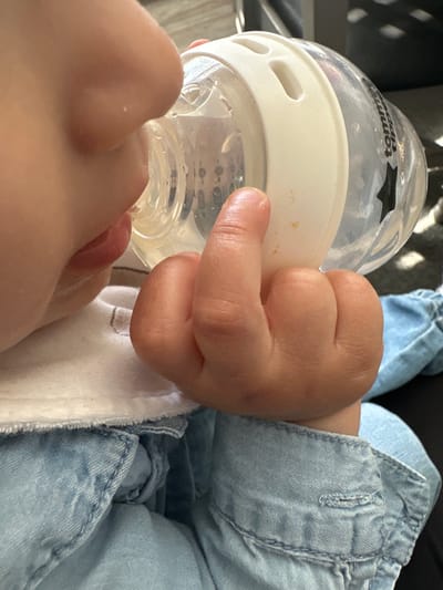 A close-up, high-angle shot captures a baby being bottle-fed in Macclesfield, United Kingdom. The baby's face is partially visible in the lower left, with their mouth latched onto the nipple of a clear baby bottle. A small, chubby hand is wrapped around the white neck ring of the bottle, holding it firmly. The brand name "Tommee Tippee" and its star logo are clearly visible on the side of the bottle.
The baby is dressed in light blue denim, possibly dungarees or an overall, showing texture, stitching, and a buttonhole detail on the fabric. A white garment, such as a babygrow or bib, is visible underneath the denim near the baby's neck. The background is softly blurred, showing dark, textured elements, suggesting an indoor setting. The lighting appears to be natural daylight. The image focuses on the intimate act of feeding, highlighting the baby's small features and grip on the bottle.