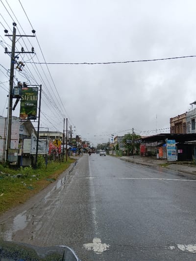A damp street stretches into the distance under a cloudy sky. Buildings line the left side of the road, interspersed with utility poles and signs. A yellow street sign and roadside vegetation are visible on the right, near a sidewalk. The road is wet and reflects light. The image is rotated and appears to be taken from inside a vehicle.