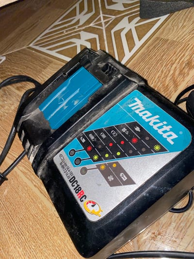 A black and turquoise Makita DC18RC battery charger is situated on a tiled floor in Baraki, Algeria. The charger features a prominent "Makita" logo and model designation "DC18RC" on its turquoise and silver-colored label. The label also specifies battery compatibility for "Li-ion:14.4-18V" and "Ni-MH:7.2-14.4V". An array of indicator lights and symbols, including those for battery charge status, error conditions, temperature, fan activity, and power, are visible on the label, with multiple red and yellow-green lights illuminated, indicating active status. The charger's black plastic casing shows visible dust and white particulate residue. A black electrical cord is connected to the charger and extends out of view to the left. The floor beneath the charger consists of light brown or tan tiles with a distinct white geometric pattern, and some areas exhibit a wood-grain texture. No individuals are present within the scene.