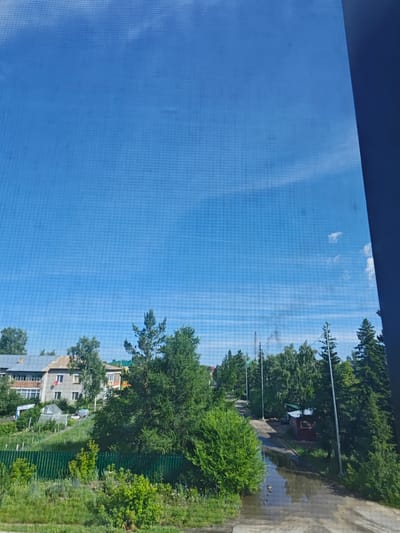 The image captures a view of a rural or semi-rural residential area, likely in a city like Оқжетпес, Kazakhstan, on a bright, clear day. The sky is a vibrant blue with some scattered, wispy white clouds. A prominent grid pattern, possibly from a screen or window, overlays the entire image, particularly noticeable in the sky.
In the foreground, a patch of green grass and various bushes and plants suggest a natural or semi-cultivated area. A green metal fence runs horizontally across the lower part of the scene. Beyond the fence, several residential buildings are visible. These appear to be multi-story houses, constructed with brick and concrete, featuring windows and balconies. Some buildings have pitched roofs. Adjacent to the houses, there are indications of gardens or cultivated plots, including what looks like a greenhouse.
A dirt or gravel road winds through the scene, leading into the distance. On the right side of the road, there is a small, red, shed-like structure with a window. Further down the road, puddles of water on the surface indicate recent rain or a drainage issue. Several tall trees, predominantly evergreens and deciduous varieties, are interspersed throughout the landscape, providing shade and a natural barrier. Power lines and utility poles are also visible in the background, suggesting the presence of infrastructure. In the far distance, there are more trees and a hint of other structures.
No people are clearly visible in the image, and no specific actions or interactions are depicted. The overall impression is one of a quiet, sunny day in a residential area with elements of both nature and human habitation.