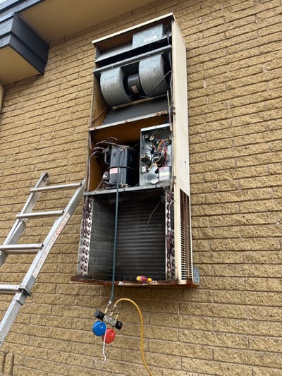 An open air conditioning unit hangs on a tan brick wall, accessed by an aluminum ladder. Inside, two cylindrical fans are visible on the left, followed by electrical components and a black motor. A large heat exchanger coil occupies the right portion. Refrigerant gauges with yellow and blue hoses are connected to the unit for servicing. Rust stains are visible along the edges of the AC unit.