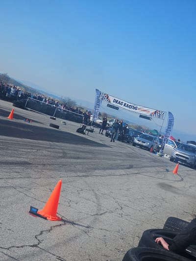 The image depicts a drag racing event in Kraynitsi, Bulgaria. A banner reading "DRAG RACING КРАИНИЦИ" spans the track. Numerous vehicles are lined up at the starting point. Spectators are visible along the sidelines behind a barrier. Orange traffic cones mark the course. The sky is clear and blue. A stack of tires is near the foreground. The asphalt surface of the track shows tire marks.