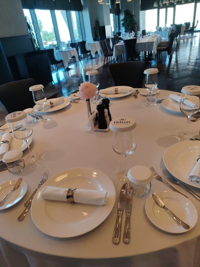 This is a medium shot of a round table set for a formal meal in what appears to be a restaurant. The table is covered with a white tablecloth. On the table are several place settings, each with a white plate, a folded white napkin secured with a decorative ring, silverware, and a wine glass. Some of the wine glasses have white paper covers with "Hilton SAMARKAND REGENCY" printed on them. There are also small glass jars with white lids on the table. A single pink flower is arranged in a small vase. In the background, through a large window, trees and buildings are visible, suggesting an outdoor view. The interior of the restaurant has a modern design with dark chairs and a polished floor. The overall impression is one of an elegant dining setting.