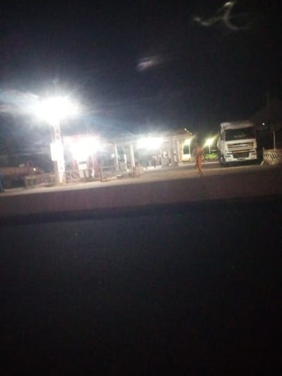 The image depicts a nighttime scene at a gas station in Baccure Kofare, Nigeria. The primary focus is on a large white truck parked at the gas station, illuminated by bright overhead lights. The gas station itself appears to be operational, with visible pumps and a covered structure.

A lone individual, seen from the back, is walking away from the gas pumps towards the right of the frame, wearing light-colored clothing that stands out against the dark surroundings. The lighting from the gas station creates a stark contrast with the dark night sky, which features some faint, wispy clouds or light reflections.

In the background, there are other structures and possibly vehicles at the gas station, but they are less distinct due to the low light conditions. The overall impression is one of a typical roadside stop during the night, with activity limited to the immediate area illuminated by the gas station's lights. The image quality is poor, with significant blur and noise, making it difficult to discern finer details. No visible text is discernible in the image.
