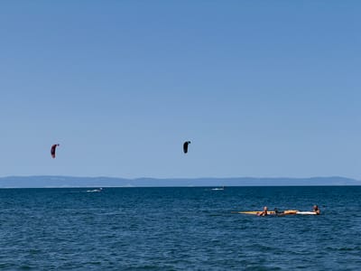 In Atiya, Bulgaria, on a clear, sunny day, two individuals are relaxing on a yellow and white inflatable raft in the calm, blue waters of the Black Sea. The raft is positioned on the right side of the frame. In the middle ground, two kite surfers are engaged in their sport, one with a red kite and the other with a black kite, both creating white wakes behind them. In the far distance, a hazy mountain range forms the coastline. The sky is a vast expanse of clear blue.