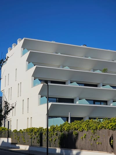 The image shows a modern white apartment building with layered balconies under a clear blue sky. The building has a geometric design with angular balconies featuring light blue glass railings. Tall, thin windows are visible on the left side of the building. A black lamppost stands in front of the building, casting a shadow on the pavement. A fence made of interwoven natural materials, covered with lush green and yellow foliage, runs along the front of the property. In the background, faint hints of mountains are visible. The scene appears to be in Deveses, Spain, given the architectural style and the presence of Mediterranean vegetation.