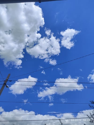 A vertical, low-angle shot captures a bright blue sky with scattered white cumulus clouds. A dark horizontal line, possibly a roof overhang, runs across the top of the frame. Several thin black wires stretch horizontally across the sky, supported by a wooden utility pole visible on the left side of the image. The pole has insulators and a lamp attached to it. In the lower right corner, a timestamp reads "17.06.2026 14:28". The scene appears to be outdoors in an urban or suburban area.