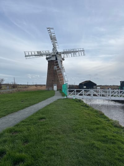 The image depicts St Benet's Abbey Windmill, located near Ludham in the North Norfolk district of the United Kingdom, under an overcast sky.

The scene features a prominent brick tower windmill with a white cap and four white, lattice-framed sails, some of which exhibit missing slats, indicating disrepair or maintenance. A white rectangular door is visible at the base of the mill, and a small white-framed window is present higher on the tower. A light grey gravel path curves from the lower-left foreground directly to the mill's entrance. Green grass covers the ground on either side of the path and along the banks of a narrow canal or river running parallel to the path on the right.

A white, metal-truss pedestrian bridge spans the canal in the mid-ground. Beyond the bridge on the far bank, a dark-colored wooden boathouse or building with a dark roof is visible, along with a partially obscured second similar structure further right. A section of green mesh fencing is positioned near the windmill's base on the right side of the door. A white and orange traffic cone stands near the canal on the right, adjacent to the bridge. In the distant left background, open fields, utility poles, and a road are discernible. No individuals are present, and no actions are observed.