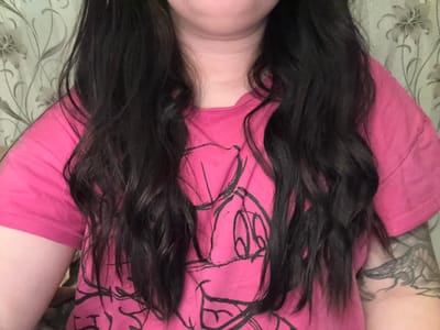 The image shows a person lying down, viewed from above. They are wearing a pink shirt with a black cartoon outline design. Long, dark, wavy hair frames the center of the image. The person's neck and chin are visible at the bottom. A tattooed arm is visible on the left side. The person is lying on a bed with a floral patterned bedspread. The location in the city of Dobrovnitsa, Bulgaria is not determinable from the visual information provided in the image.