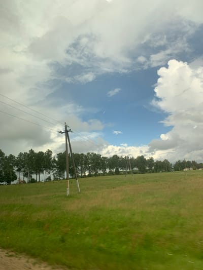 The image depicts a rural landscape under a partly cloudy sky, likely in or near Юнькаўскі сельскі Савет, Belarus. In the foreground, a lush green field with some patches of drier grass stretches across the frame. A dirt road or path is visible at the very bottom left corner.
Dominating the mid-ground is a row of deciduous trees, their foliage a vibrant green, lining the crest of a gentle hill. A prominent wooden utility pole with multiple crossbars and wires stretches across the scene from left to right, positioned in front of the tree line. Another similar pole is visible further in the distance to the right. In the far distance, a few scattered buildings with light-colored roofs can be discerned, hinting at a small settlement.
The sky above is a dynamic mix of bright blue patches and large, billowing white and gray clouds, suggesting a transition in weather. The overall impression is one of an open, somewhat windswept countryside. No people or animals are visible in the image.