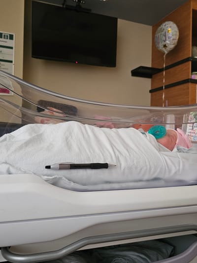 This image depicts a peaceful scene of a newborn baby resting in a hospital bassinet. The baby, swaddled in a soft white blanket and wearing a light pink hat, has a blue-green pacifier in its mouth and appears to be sleeping soundly. A black pen is tucked along the inside edge of the bassinet, close to the baby.

The bassinet features clear acrylic sides and a white frame, characteristic of hospital maternity wards. In the background, a white Mylar balloon with "It's a Girl!" text and star designs is visible, floating near a wooden wall-mounted shelf. Below the shelf, a dark flat-screen television is mounted on the wall, showing a small red indicator light. Medical documents or charts with green and white branding can be seen on the wall beneath the television. The room has light-colored walls and appears brightly lit, suggesting a daytime setting or well-illuminated interior. The overall atmosphere conveys new life and quiet tenderness. The image itself is captured with a sideways orientation, making the background elements appear vertical.