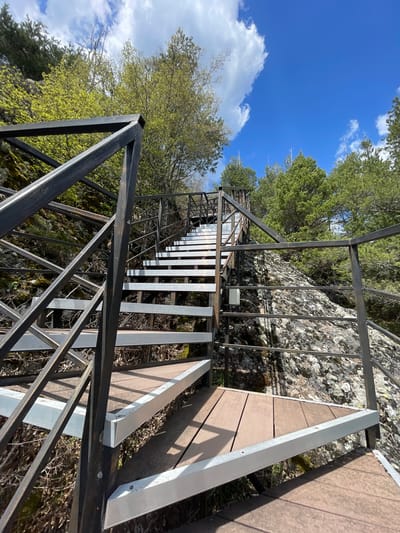 A low-angle shot depicts metal stairs with wooden landings ascending a rocky terrain. The stairs feature a dark metal frame and light metal steps. A railing encloses the staircase. Vegetation, including trees, is visible along the sides of the stairs. The sky is blue with scattered clouds. The location is Katranica, Bulgaria.