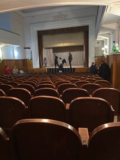 The image depicts the interior of a theater or auditorium, likely in Voiniagovo, Bulgaria. Rows of dark wooden seats occupy the foreground, leading to a stage at the far end. Several people stand on the stage, possibly performing or rehearsing. The stage is framed by a decorative archway and curtains. Other people are seated or standing in the aisles. Green and white balloons decorate the sides of the room, suggesting a celebratory event.