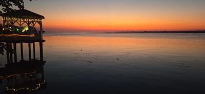 A photograph captures a sunset over Lake Constance in Bregenz, Austria. In the foreground, a wooden gazebo on stilts stands in the water, adorned with string lights. Its reflection is visible on the calm surface of the lake. The sky displays a gradient of colors, transitioning from a soft blue at the top to vibrant orange and red hues near the horizon. In the distance, a silhouetted shoreline with undulating hills marks the edge of the lake. The water is tranquil, with subtle ripples and a scattering of organic debris on its surface.
