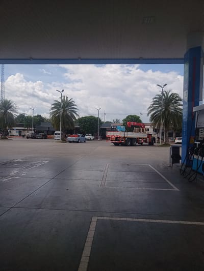 The image shows a view from under the canopy of a gas station in Tha Ruea, Thailand. The forecourt is paved with large concrete slabs marked with thin lines. In the distance, several cars are parked. A large truck with a crane is parked further away. Palm trees are visible on either side of the scene. Above the scene, a cloudy sky is visible. The gas station has blue support pillars with gas pumps attached. A trash can is positioned next to one of the pillars.