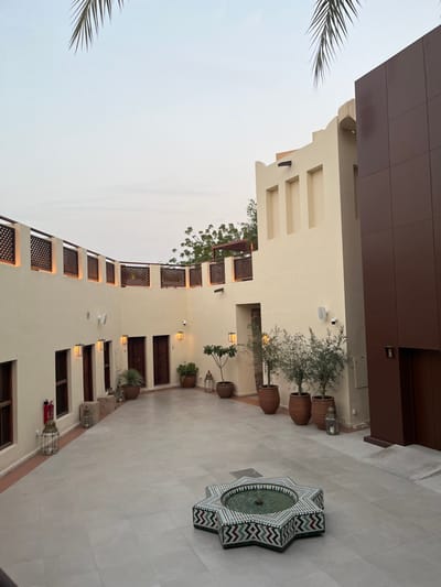This image depicts an empty, traditional-style courtyard within a building complex in Sharjah, United Arab Emirates, likely a heritage area or cultural space. The scene is illuminated by ambient light, suggesting dusk or dawn under an overcast sky, with a partial view of a palm frond in the upper frame.
The courtyard is enclosed by pale cream-colored stucco buildings with smooth finishes. The ground is paved with light grey tiles. In the center of the courtyard, an eight-pointed star-shaped fountain is visible, featuring a basin tiled in green and an exterior adorned with geometric patterns in green, white, and a dark reddish-brown.
The left and back walls of the courtyard feature multiple rectangular wooden doors and recessed windows with dark frames, characteristic of traditional Emirati architecture. Several decorative wall-mounted lanterns emit a warm light. Along the base of the walls, numerous potted plants of varying sizes, including several small trees, are arranged in terracotta pots. Additional traditional-style metal lanterns rest on the ground. A red fire extinguisher is mounted on the wall to the left. The upper section of the walls is topped with crenellated-style elements and a series of dark wooden lattice screens, illuminated from below.
To the right, a multi-story section of the building includes a contemporary dark brown paneled facade contrasting with the traditional cream structure. This rightmost building section also features a cream-colored wall with three prominent, recessed rectangular cutouts at its upper level. White wall-mounted security cameras and speakers are visible throughout the courtyard. No individuals are present in the scene.