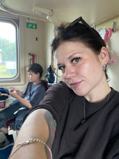 A woman with dark hair, wearing a dark brown shirt and a silver necklace, takes a selfie on a train. She has sunglasses perched on her head and several ear piercings. Her left arm, adorned with a tattoo, is visible in the foreground. In the background, a young boy with brown hair, wearing a dark gray t-shirt, is seated and looking at his phone. He is wearing dark pants and sneakers. The train car has a window with a view of trees and buildings, and there are various signs and safety equipment visible on the wall. The location is identified as Ershovka, Russia.