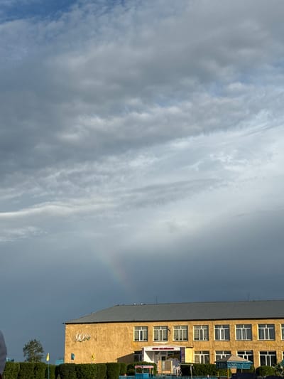 The image shows a building with a yellow facade and a gray roof in what appears to be a school or community center in the city of Өндіріс, Kazakhstan. The building has multiple windows and an entrance. There is a sign above the entrance that reads "90 ЖЫЛ" with a floral design. In the background, a faint rainbow is visible against a cloudy sky, with dark storm clouds dominating the upper portion of the frame. The foreground features a neatly trimmed hedge and some flags. The overall scene suggests an outdoor setting in a populated area.