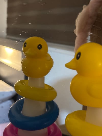 This image shows a close-up, slightly distorted view of yellow rubber ducks and colorful stacking rings, likely part of a bath toy set. The scene appears to be set in a bathroom, with reflections visible on a shiny surface, possibly a bathtub or a wet countertop. The lighting suggests an indoor setting, with a bright light source reflecting prominently in the lower part of the image. There are small water droplets or bubbles scattered across the reflective surface and on the toys themselves, indicating they are either wet or in a damp environment. The composition is close-up, focusing on the textures and colors of the toys. The orientation of the image is slightly tilted. There is no visible text. The overall impression is one of playful, bath time context, possibly with children's toys.