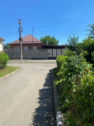 A paved road curves into the foreground, leading towards a long, decorative concrete fence with arched details. Behind the fence stands a two-story brick house with a reddish-brown tiled roof. A tall utility pole with numerous wires and a light fixture is situated in front of the house. To the right of the road, a dense green hedge lines the curb, casting shadows onto the pavement. Beyond the fence, a wide, light-colored metal gate is visible. The sky is a clear, bright blue. The scene appears to be a residential area in the city of Zapadny, Russia.