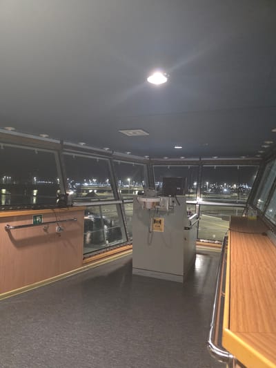 The image shows the interior of a ship's bridge at night, overlooking a brightly lit port. The bridge is equipped with large windows that offer a panoramic view of the port facilities, including numerous lights, cranes, and water. Inside the bridge, there is a console with electronic equipment and a computer monitor. A railing runs along the side, and wooden cabinetry is visible. The floor is dark gray and appears to be made of linoleum. There are no people visible in the image. The overall impression is one of a functional, operational environment within a vessel docked at a port.