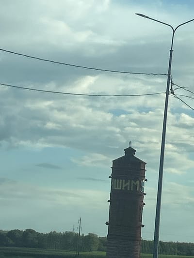 The image shows a water tower with the Russian word "ШИМ" painted on it in yellow letters. The water tower is made of dark brick and has a conical roof. It is situated in a rural setting, with trees and fields visible in the background. Power lines and a lamppost are also present in the foreground, suggesting an outdoor scene along a road. The sky is cloudy, with patches of blue sky visible. The overall lighting suggests it is daytime, possibly late afternoon or early morning due to the soft light. There are no people visible in the image. The word "ШИМ" could refer to a place name, an organization, or a brand.