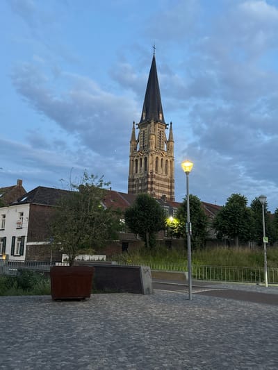 In Sittard, Netherlands, at dusk, a church tower with a pointed spire dominates the skyline. The tower, constructed from light-colored stone with dark horizontal stripes, features arched windows and decorative elements. In front of the tower, several buildings are visible, including a white house with green shutters and a red-tiled roof, and adjacent brick structures. A small tree stands in a large, weathered metal planter in the foreground. The ground is paved with cobblestones. Two lampposts illuminate the area, casting a warm glow. The sky is a pale blue with scattered clouds, and the overall atmosphere suggests an early evening in a European town.