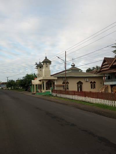 A mosque with two minarets is visible from the side of a paved road in Tocina, Indonesia. The mosque is light-colored with green accents and a green roof. The main building of the mosque has a dome and a sloped metal roof. A white and brown wooden fence runs along the side of the road, separating it from the mosque's property. Trees and other buildings are visible in the background. The sky is cloudy and light blue.