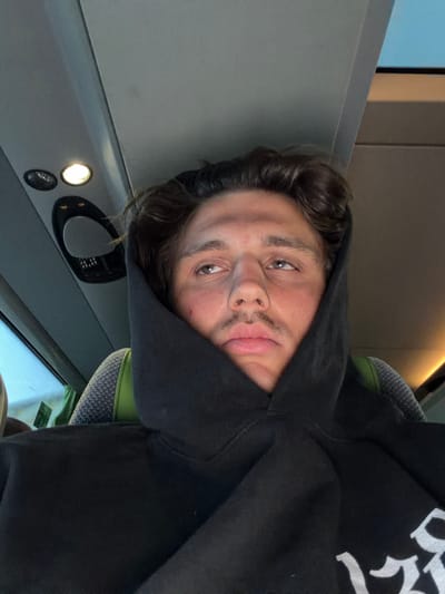 A close-up, rotated portrait shows a young person wearing a black hooded sweatshirt. The hood is pulled up, obscuring part of their face and creating a shadow. Their left eye is visible, looking directly at the camera with a neutral expression. They have dark, curly hair and a faint mustache. The background appears to be the interior of a vehicle, possibly a train or bus, with gray paneling and illuminated lights. A patterned seat is visible to the right of the person. The image seems to have been taken indoors during daylight hours, as indicated by the ambient light and the subject's attire. The overall mood is casual and introspective. No text is visible in the image. The location context is Künersberg, Germany.